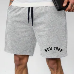 A man wearing a pair of heather grey men's sweat shorts with a 'New York 1998' graphic.